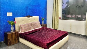 Une chambre avec un mur bleu à texture, un lit recouvert d’une couverture rouge, deux tables de chevet ornées de vases et une fenêtre donnant sur des palmiers.