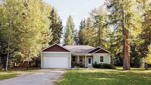 A single-story house with a garage, surrounded by tall trees and a well-maintained lawn. - Charming three bedroom near Glacier National Park (Columbia Falls)