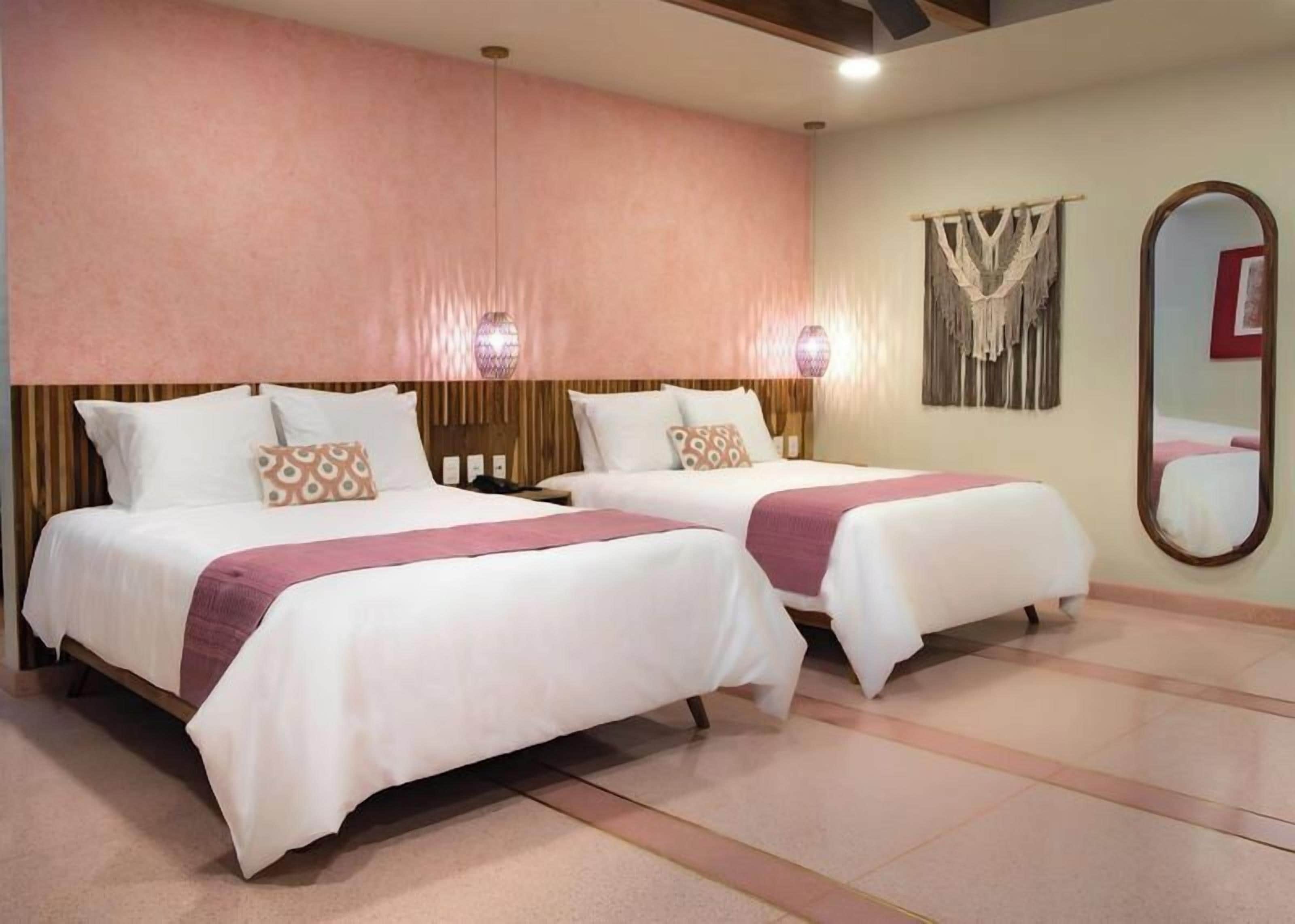 A hotel room with two beds, each with white bedding and a pink throw. There are wall-mounted lights and a large mirror on the wall.