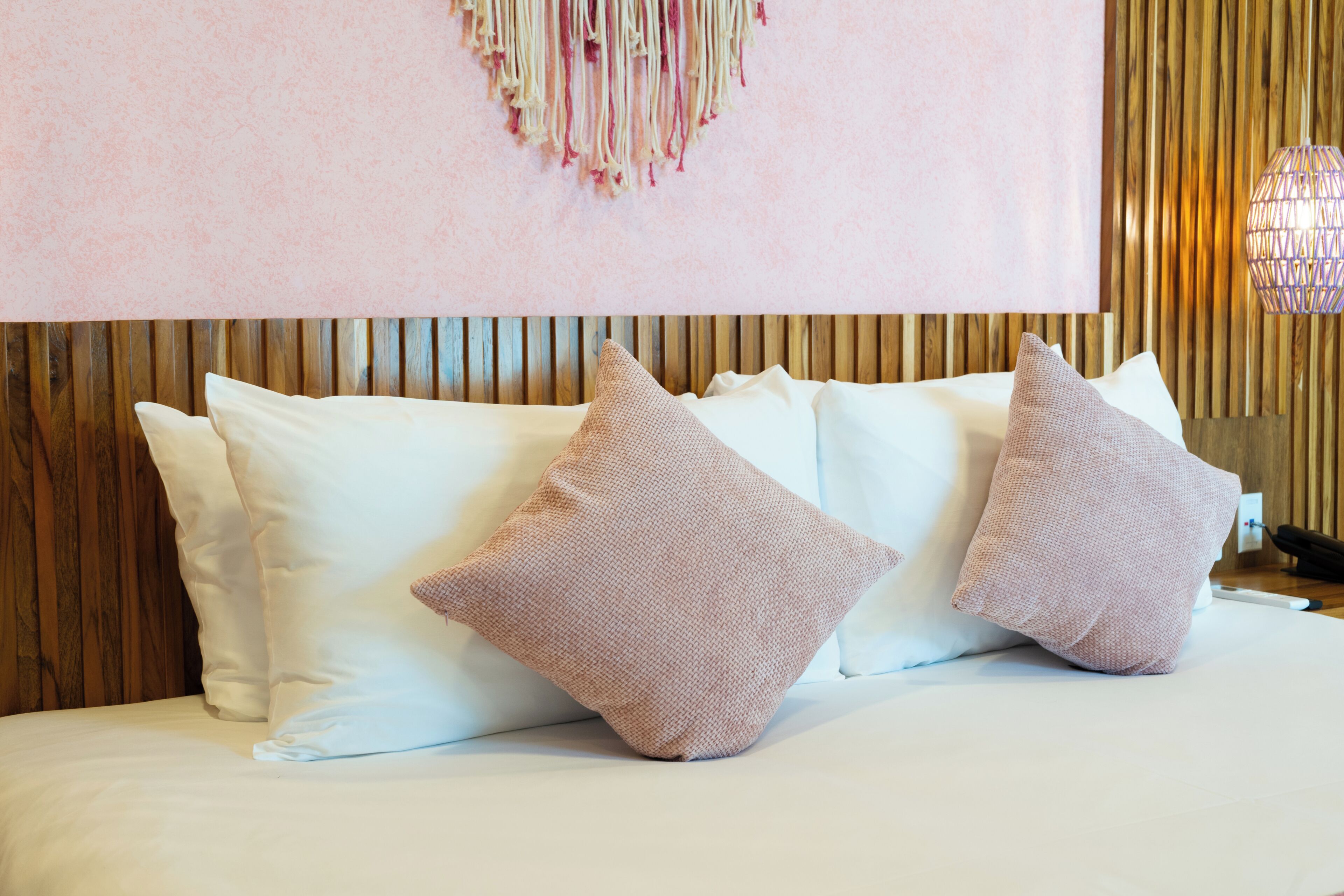 A bed with white bedding and two pink pillows, a wooden headboard, and a hanging light fixture.