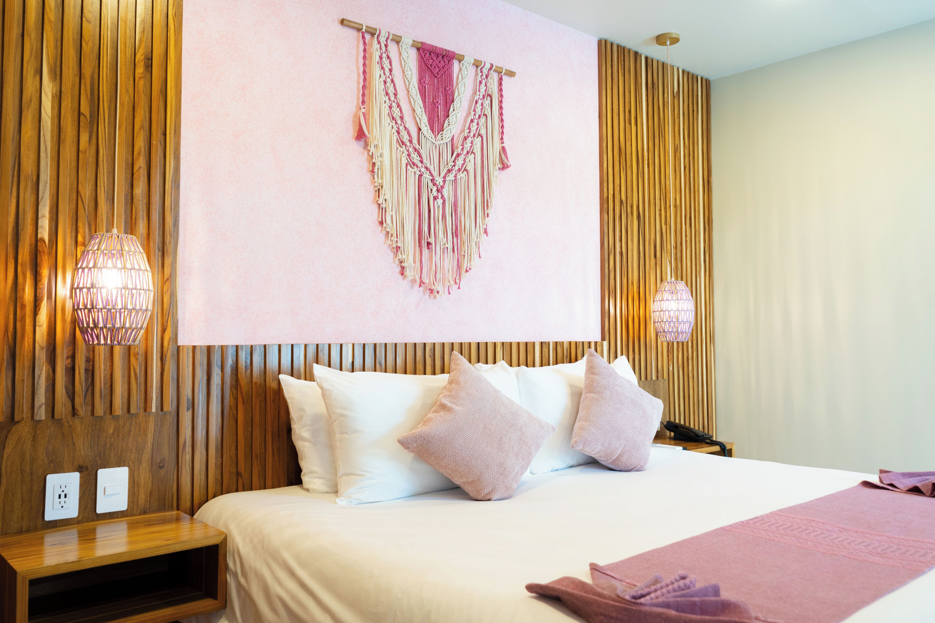 Ein Hotelzimmer mit Holz-Kopfteil, einem Bett mit weißen Bettwäsche und rosafarbenen Kissen sowie einer Wanddekoration aus Makramee.