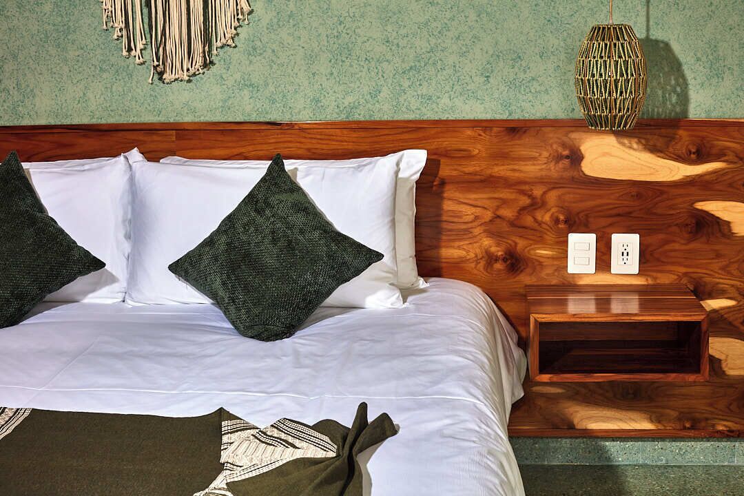 A neatly made bed with white linens and two dark green pillows, a wooden nightstand with two electrical outlets, and a woven wall hanging above the bed.