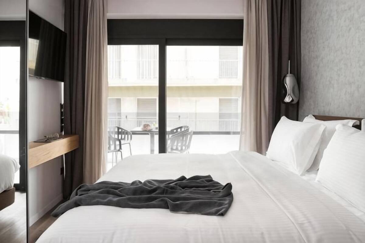 A hotel room with a bed, a window with curtains, a balcony with chairs, and a wall-mounted TV.