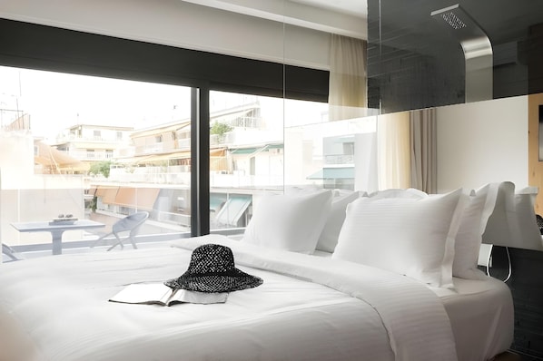 Una habitación de hotel moderna con una cama grande, ropa de cama blanca, un sombrero negro y un libro. La habitación tiene un ventanal con vistas a edificios.