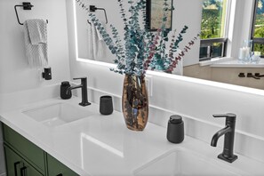 Un baño moderno con un lavamanos blanco, grifos negros y un florero con follaje verde y rojo.