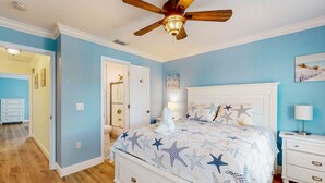 Quarto com cama, mesas de cabeceira, ventilador de teto e porta do banheiro.