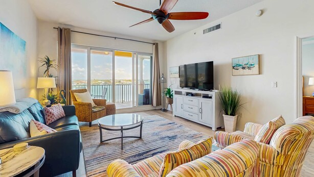 A spacious living room with a ceiling fan, a large TV, a balcony with a view, and a striped sofa. - Dog friendly bayfront 4th floor condo with bay views heated pool and spa WD (Clearwater)