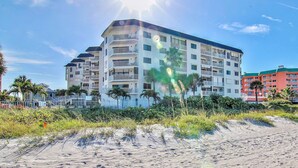 A multi-story hotel with balconies, located near a sandy beach with palm trees.