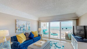 A living room with a blue sofa, a coffee table, and a TV. There is a balcony with a view of the ocean. - Chic and comfortable Gulf front condo snowbird and dog friendly pool beaches (Belleair Beach)
