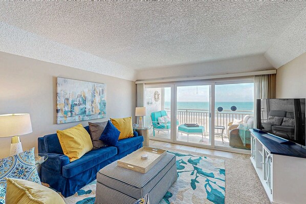 A living room with a blue sofa, a coffee table, and a TV. There is a balcony with a view of the ocean. - Chic and comfortable Gulf front condo snowbird and dog friendly pool beaches (Belleair Beach)