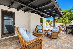 A covered patio area with wicker furniture, a glass table, and a view of a swimming pool and palm trees.