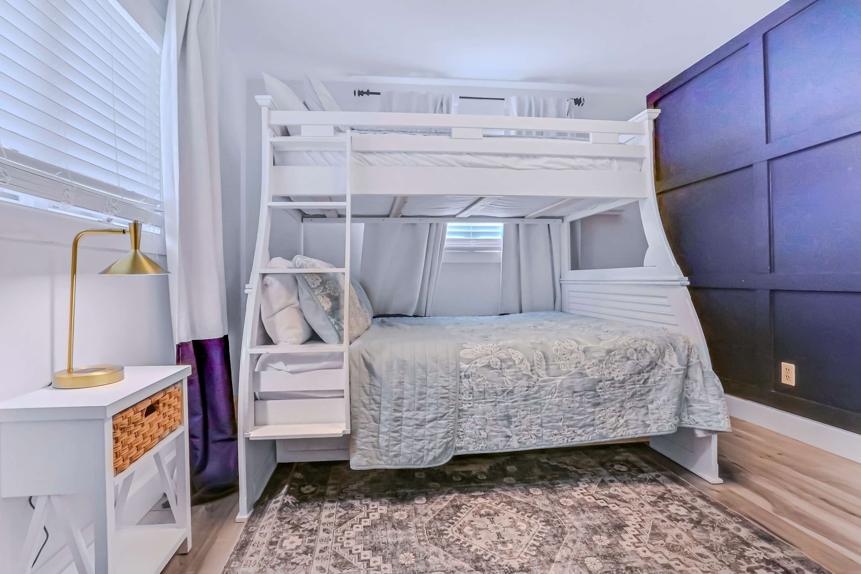 Een stapelbed met een lichtblauwe sprei, een witte nachtkastje met een gouden lamp, en een vloerkleed met een patroon.