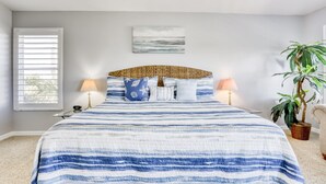 Un dormitorio con una cama, dos lámparas de noche, una mesita de noche, un reloj de pared, una ventana con persianas, una obra de arte colgada en la pared y una planta en maceta.