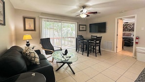 A living room with a black sofa, a glass coffee table, a dining table with chairs, a ceiling fan, and a TV mounted on the wall. - Centrally located condo with heated pool balcony washer dryer and central AC (Clearwater)