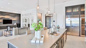 A modern kitchen with a large island, stainless steel appliances, pendant lighting, and a seating area with a TV.