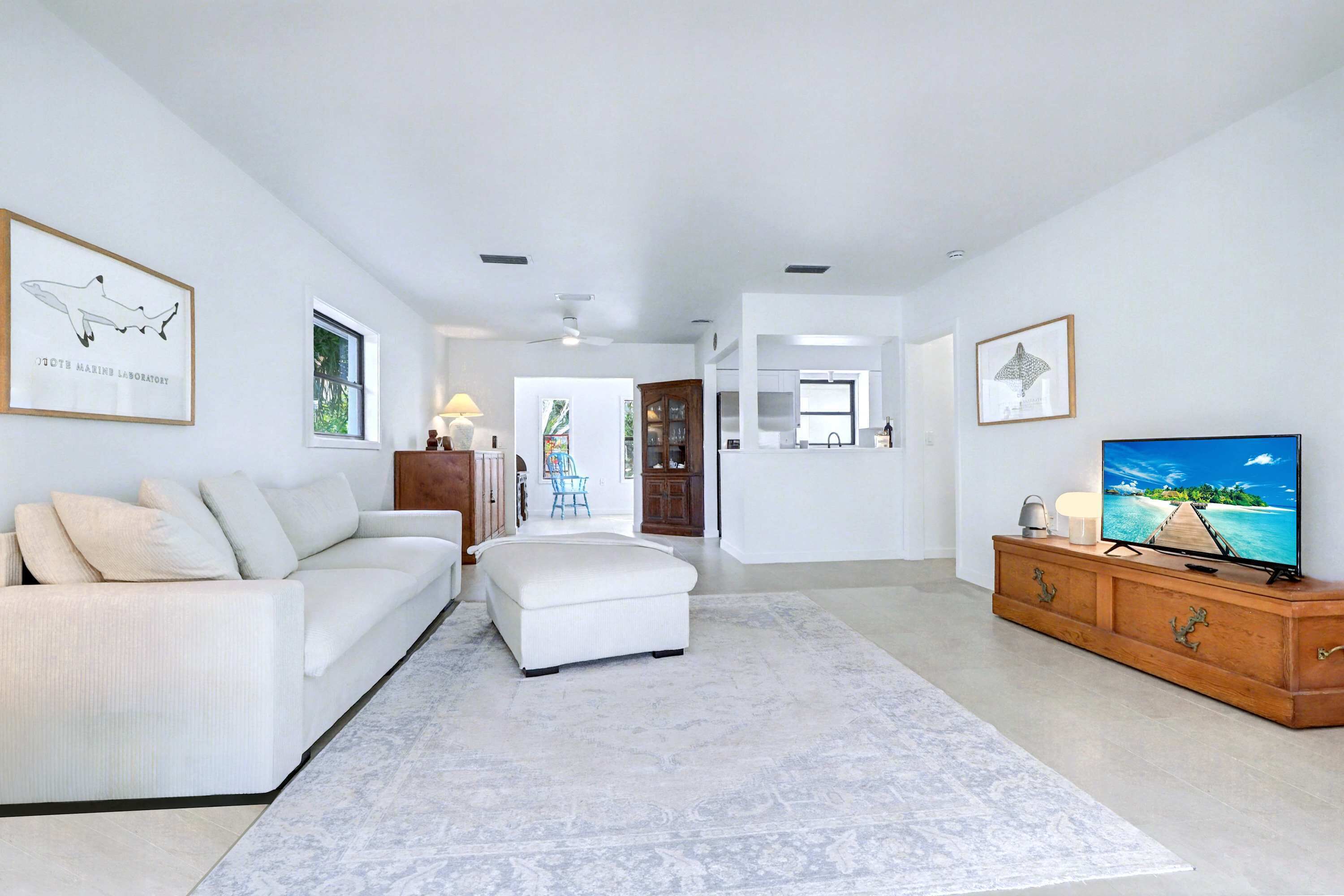 A spacious living room with a white sofa, a white ottoman, a wooden TV stand, and a large flat-screen TV.