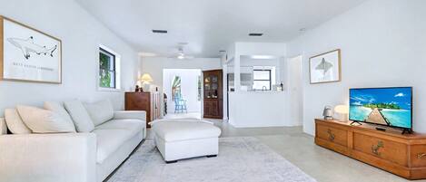A spacious living room with a white sofa, a white ottoman, a wooden TV stand, and a large flat-screen TV.