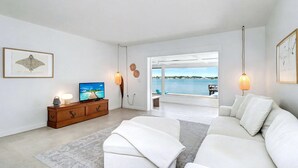 Una sala de estar moderna con un sofá blanco grande, un televisor sobre un mueble de madera y vistas al mar a través de ventanas amplias.