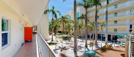 Pasillo de hotel con zona de piscina, palmeras y mobiliario exterior.