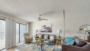 Ein geräumiges Wohnzimmer mit Deckenventilator, einer Couch, einem Couchtisch und einem großen Fenster mit Vorhängen.