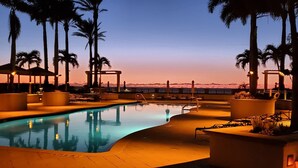 Zona de piscina con sillones de descanso y palmeras, rodeada por un muro bajo y vista al atardecer.