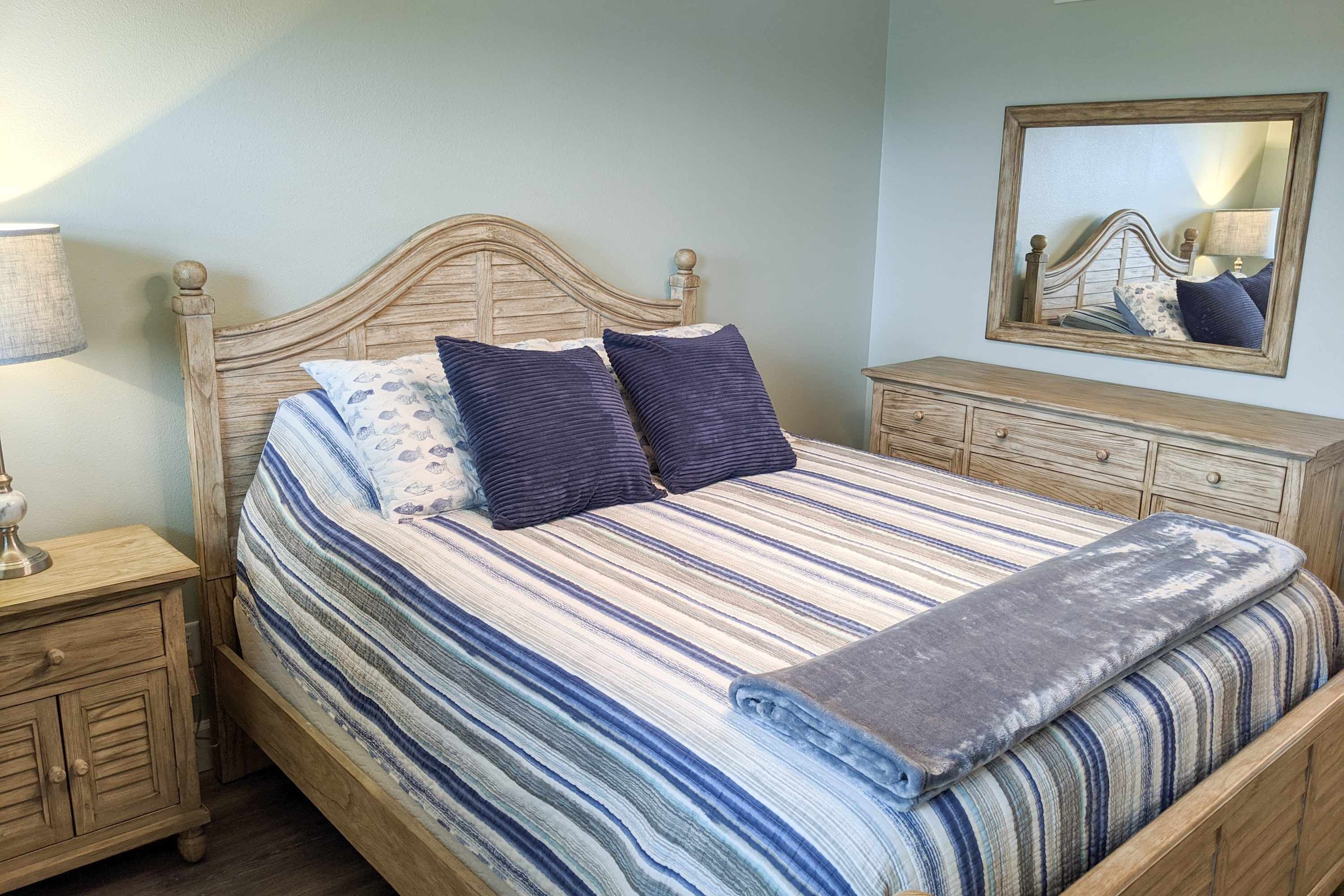 Un lit bien fait, avec une couvre-lit à rayures, deux oreillers et une couverture. On y trouve une table de chevet en bois avec une lampe, ainsi qu’une commode avec des tiroirs. Un miroir est accroché au mur, au-dessus de la commode.
