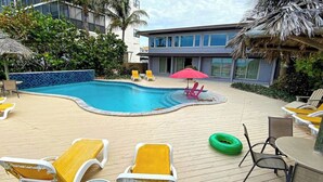Un área de piscina con sillas de descanso y una mesa, rodeada por una terraza de madera y palmeras.