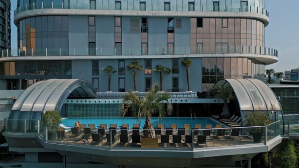 Un hotel moderno con fachada de vidrio curva, una piscina en la azotea con tumbonas y vistas de la ciudad circundante.
