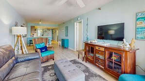 Een woonkamer met een grijze bank, een blauwe fauteuil, een houten tv-meubel en een plafondventilator.