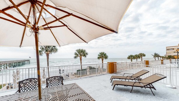 Un patio frente a la playa con mesa y sillas, un gran paraguas y sillones de descanso.