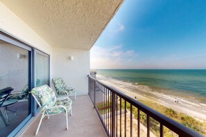 A balcony with a view of the beach, a seating area with chairs, and a glass door. - Madeira Siesta Condo in Madeira Beach (Madeira Beach)