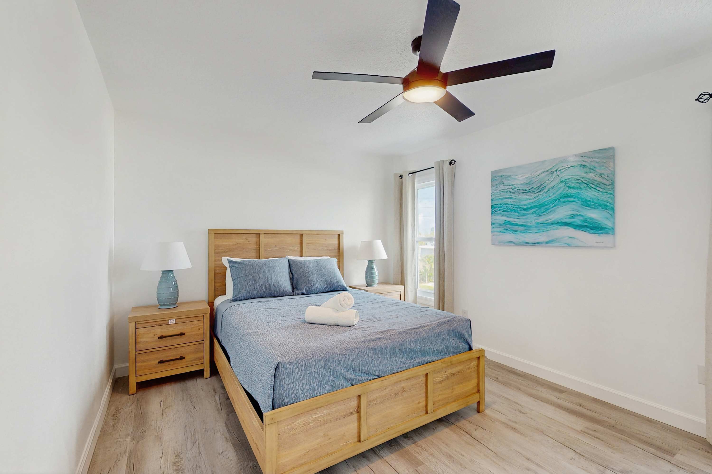 A bedroom with a wooden bed, nightstands, a ceiling fan, a window with curtains, and a wall painting.