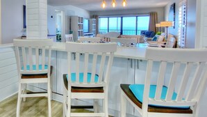 Een moderne keuken met witte barkrukken, een eettafel en uitzicht op de oceaan door grote ramen.