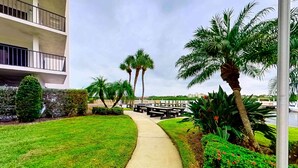 A pathway leading to a building with a balcony, surrounded by well-maintained grass and palm trees. - Coastal condo with beach and pool access balcony WD central air and water views (Indian Shores)