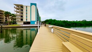 A wooden boardwalk leading to a building with multiple balconies, a pool, and a view of a green landscape. - Coastal condo with beach and pool access balcony WD central air and water views (Indian Shores)