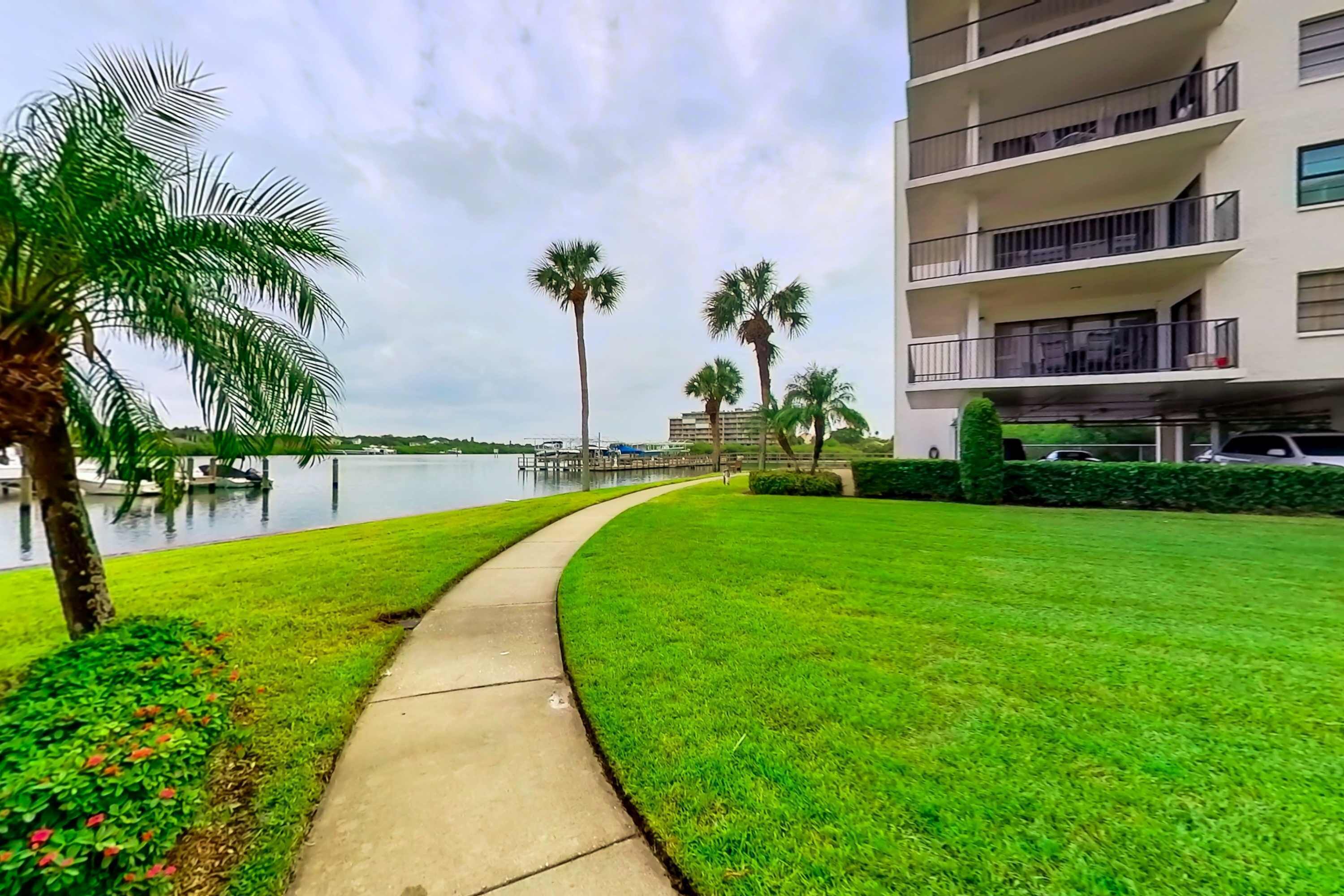 A well-maintained grassy area with a concrete pathway, palm trees, and a body of water in the background.