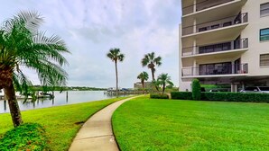 A well-maintained grassy area with a concrete pathway, palm trees, and a body of water in the background. - Coastal condo with beach and pool access balcony WD central air and water views (Indian Shores)
