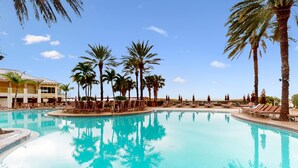 Una piscina grande y cristalina rodeada de palmeras y sillas de descanso, con un edificio al fondo.