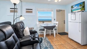 A compact living space with a sofa, a dining table with chairs, a ceiling fan, a refrigerator, and a view of the ocean through a window.