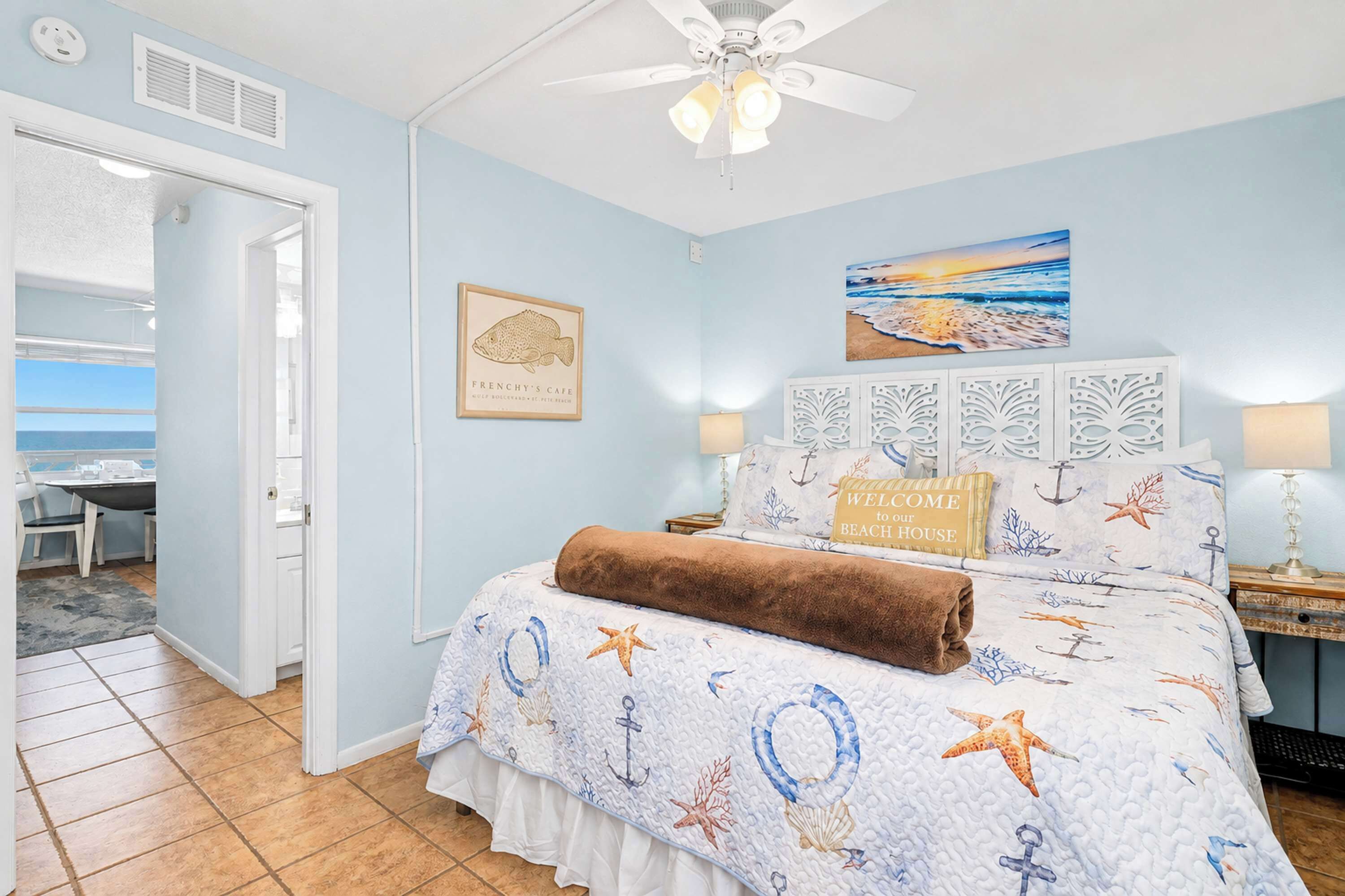 Une chambre à coucher avec un lit, un ventilateur de plafond, un tableau représentant une plage et une photo de poisson encadrée.