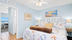 A bedroom with a bed, a ceiling fan, a painting of a beach, and a framed fish picture.