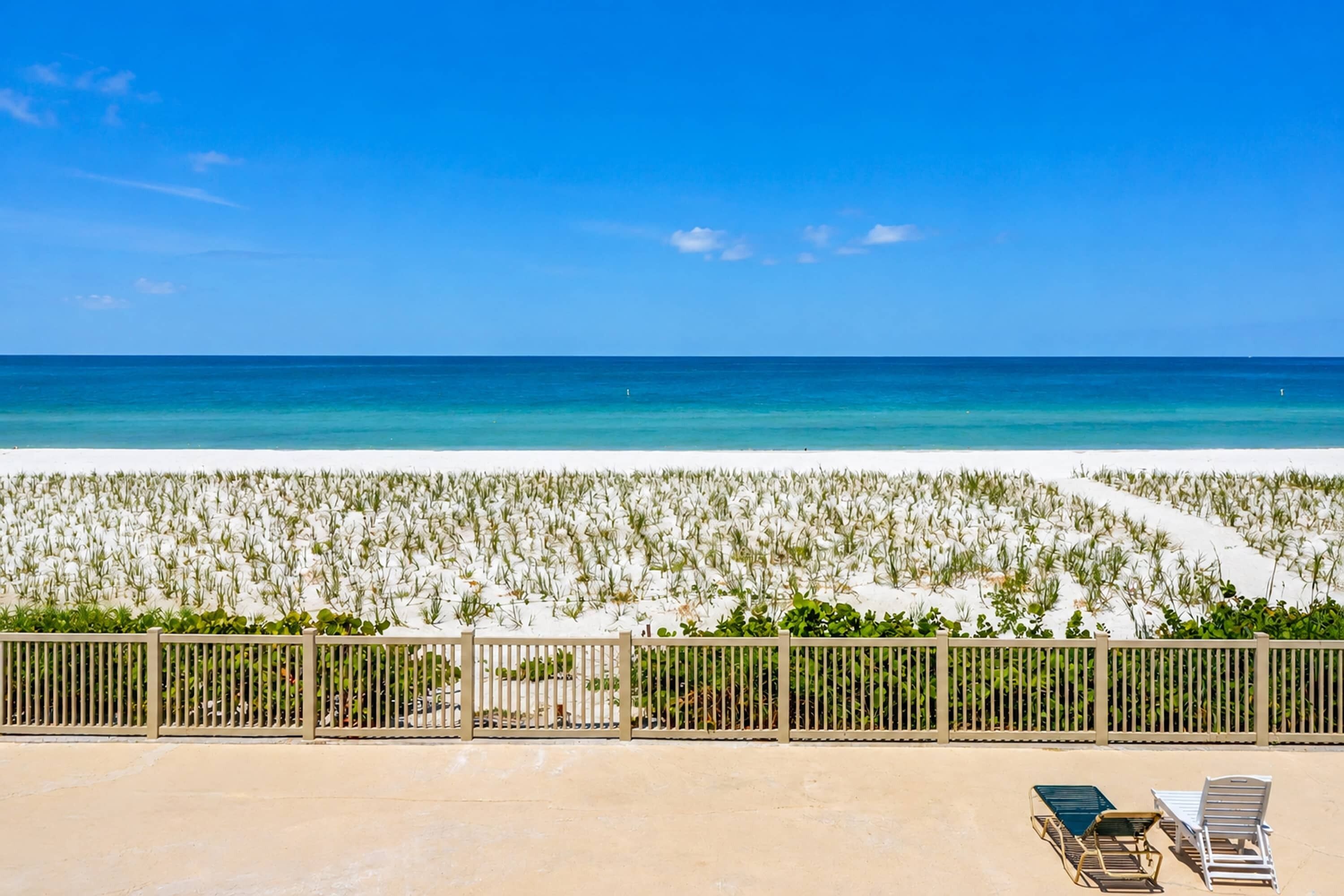 Een strand met helder uitzicht op de oceaan, een zandgebied met begroeiing, en een omheind gebied met ligstoelen.
