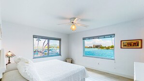 Een slaapkamer met een bed, twee ramen met uitzicht op een strand en stadsbeeld, een plafondventilator en een kleine tafel met een lamp.