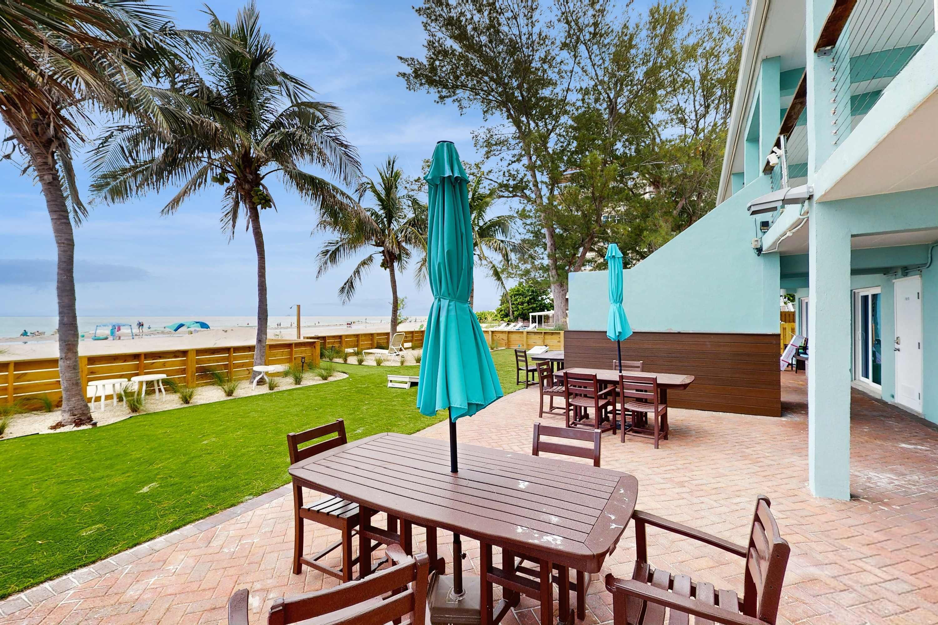 Un patio exterior con mesas y sillas de madera, césped verde y vistas a la playa y palmeras.