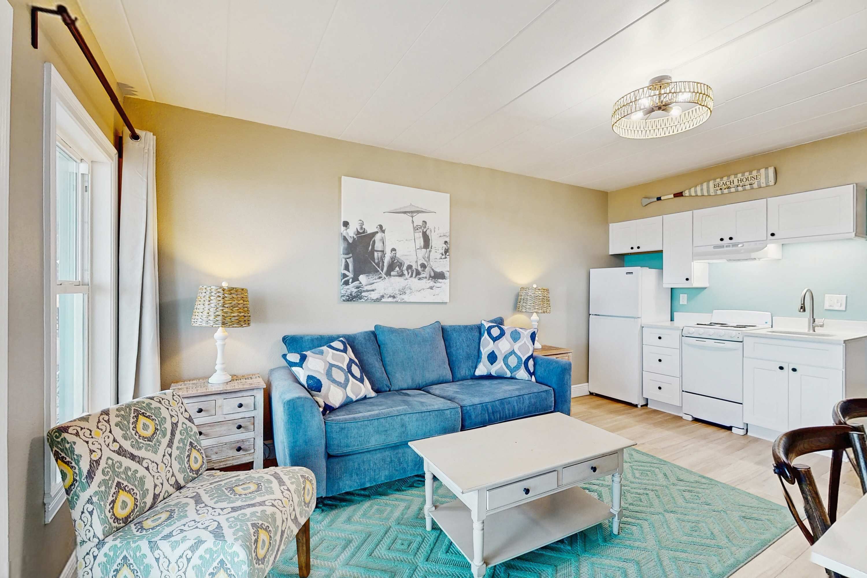 Ein Wohnzimmer mit blauem Sofa, einem gemusterten Sessel und einem weißen Couchtisch. Es gibt eine Küchenzeile mit weißen Schränken und einem Kühlschrank. Ein Wandbehang im Strandhausstil ist sichtbar.