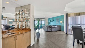 Una cocina moderna con encimeras de granito, gabinetes de madera y una isla integrada con pileta. Hay un comedor con una mesa y sillas grises, y una sala de estar con una pared de acento azul y un ventanal grande.
