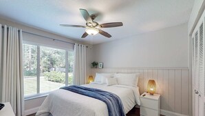 Ein Schlafzimmer mit Bett, Nachttischlampen, Deckenventilator, einem Fenster mit Vorhängen und Blick ins Grüne.