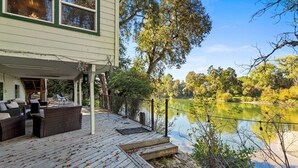 Een houten terras met buitenmeubilair, met uitzicht op een meer met bomen en een gebouw op de achtergrond.