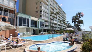 Zona de piscina de hotel con sillones reclinables, una bañera de hidromasaje y un edificio grande al fondo.