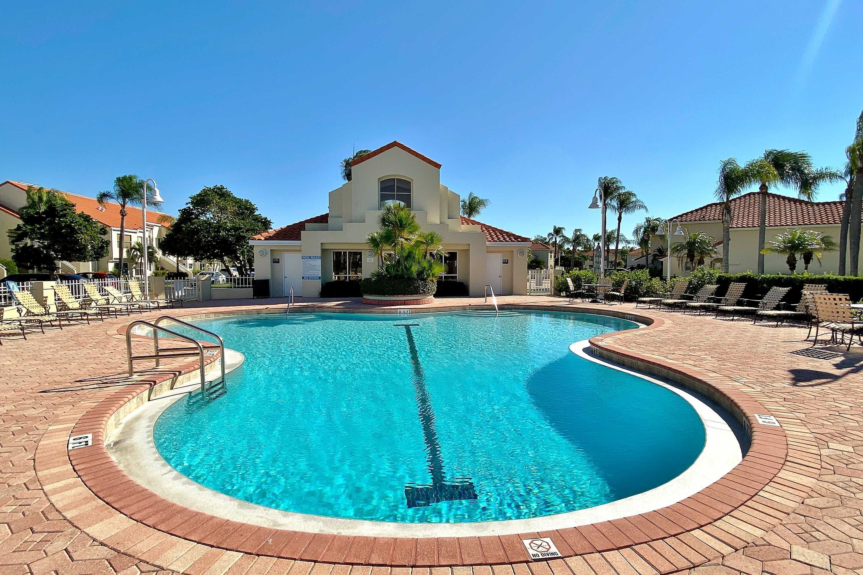 A swimming pool with a surrounding brick deck, a building with a red-tiled roof, and palm trees.